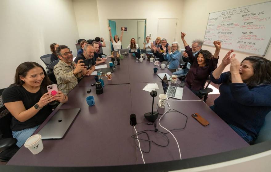 A boardroom table full of people celebrates at Pulitzer Prize win, taking selfies, raising arms, shouting, etc.