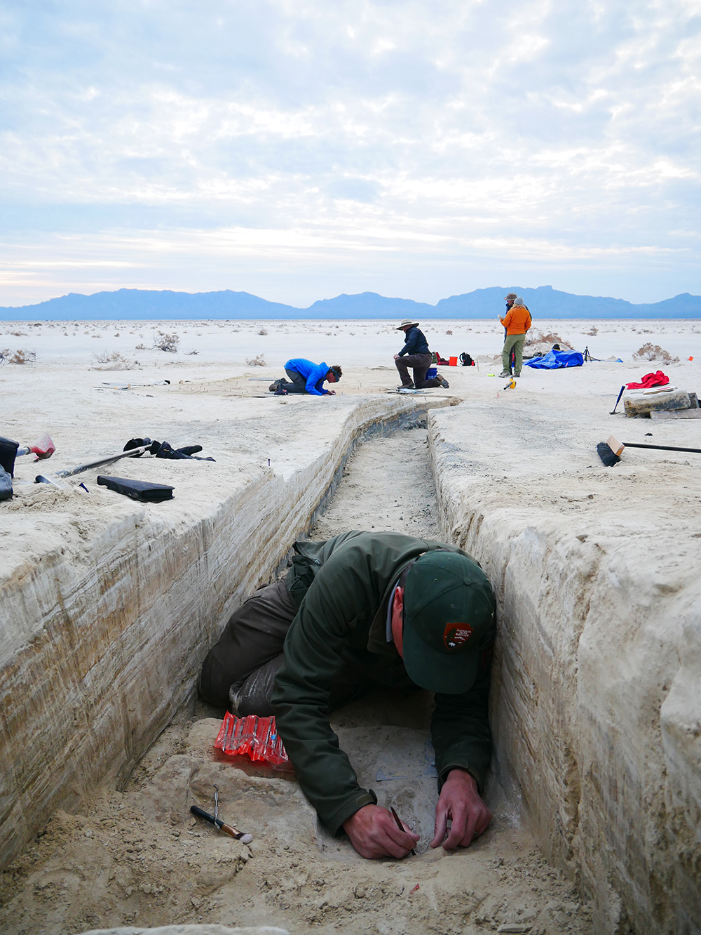 A man wearing a National Park Service uniform sifts through a trench dug in a white sand landscape, with 3 other researchers in the background.