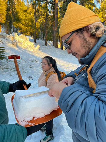 A student in a hat and warm jacket measuring snowpack on a snowy, forested slope