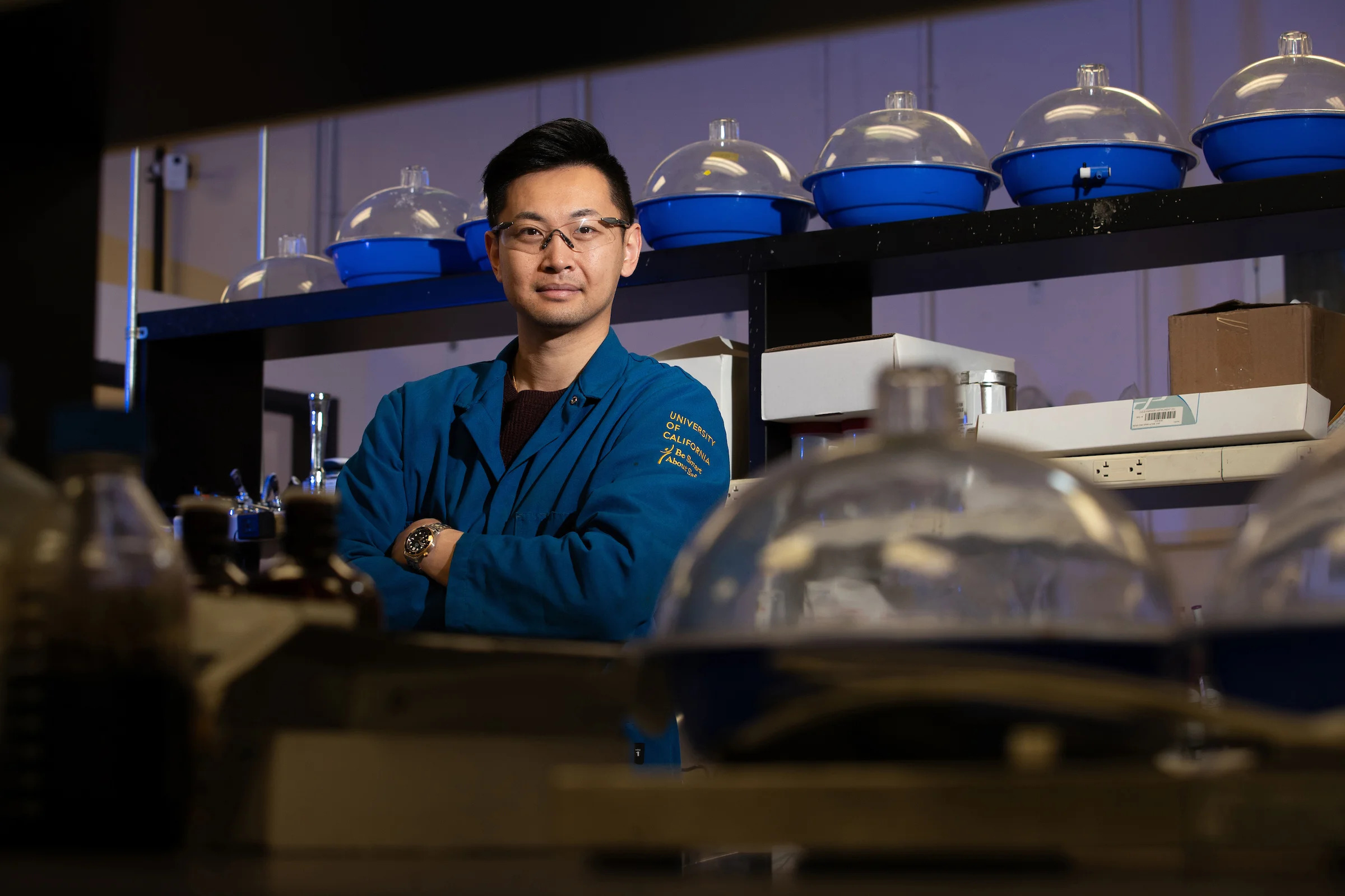 A young Asian researcher in a lab in a blue lab coat, arms crossed