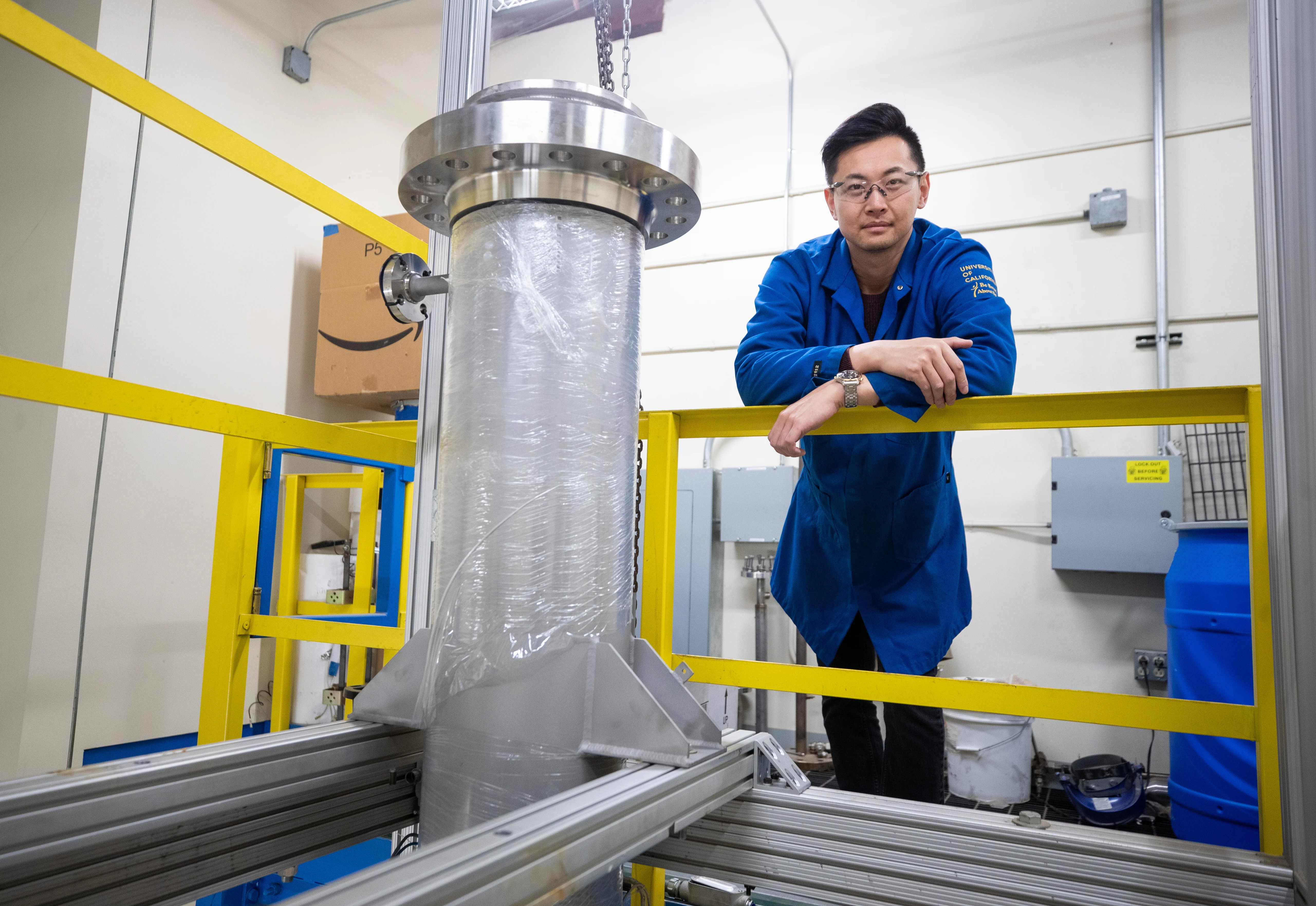 A young Asian male researcher in a blue lab jacket next to a 20-gallon CELF reactor indoors