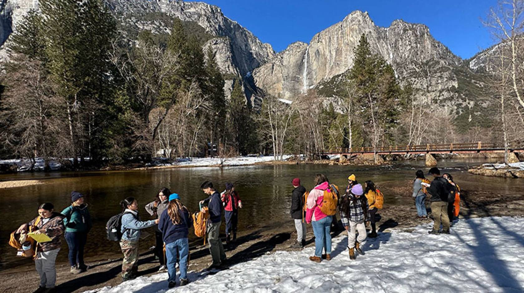 On a sunny day, a group of students gathers on the banks of a river, with snow on the ground and dramatic mountain peaks with a waterfall and forest in the background. 