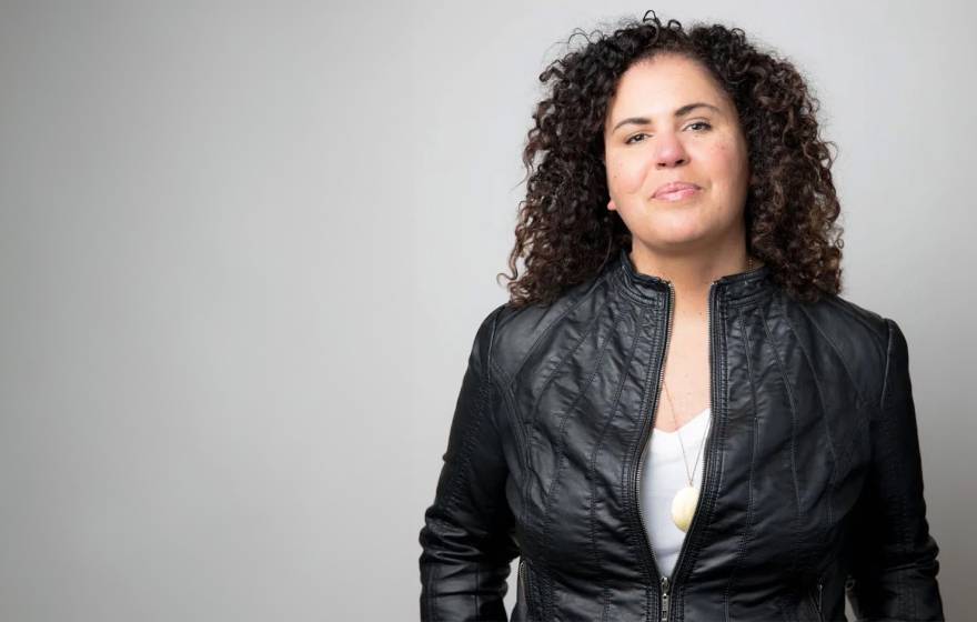 Safiya Noble, wearing a cool black leather jacket, looks resolute in a waist-up portrait against a gray background