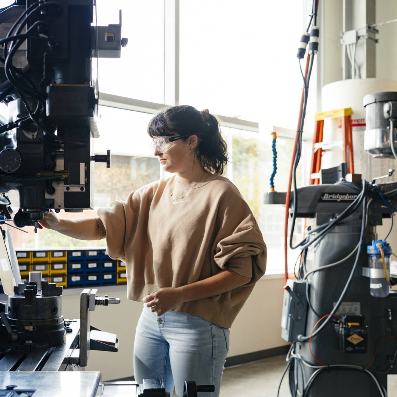A woman in jeans and a brown sweater wearing safety glasses uses a large machine in a lab lit by natural light from a large wall of windows