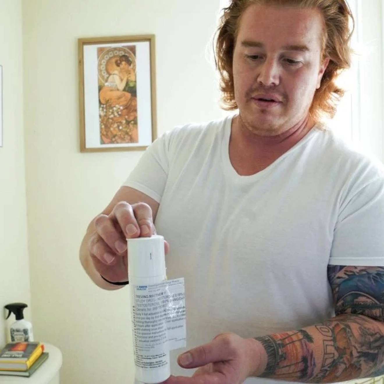 A man with arm tattoos in a white t-shirt holds a white plastic bottle, standing in a well lit room with white walls.