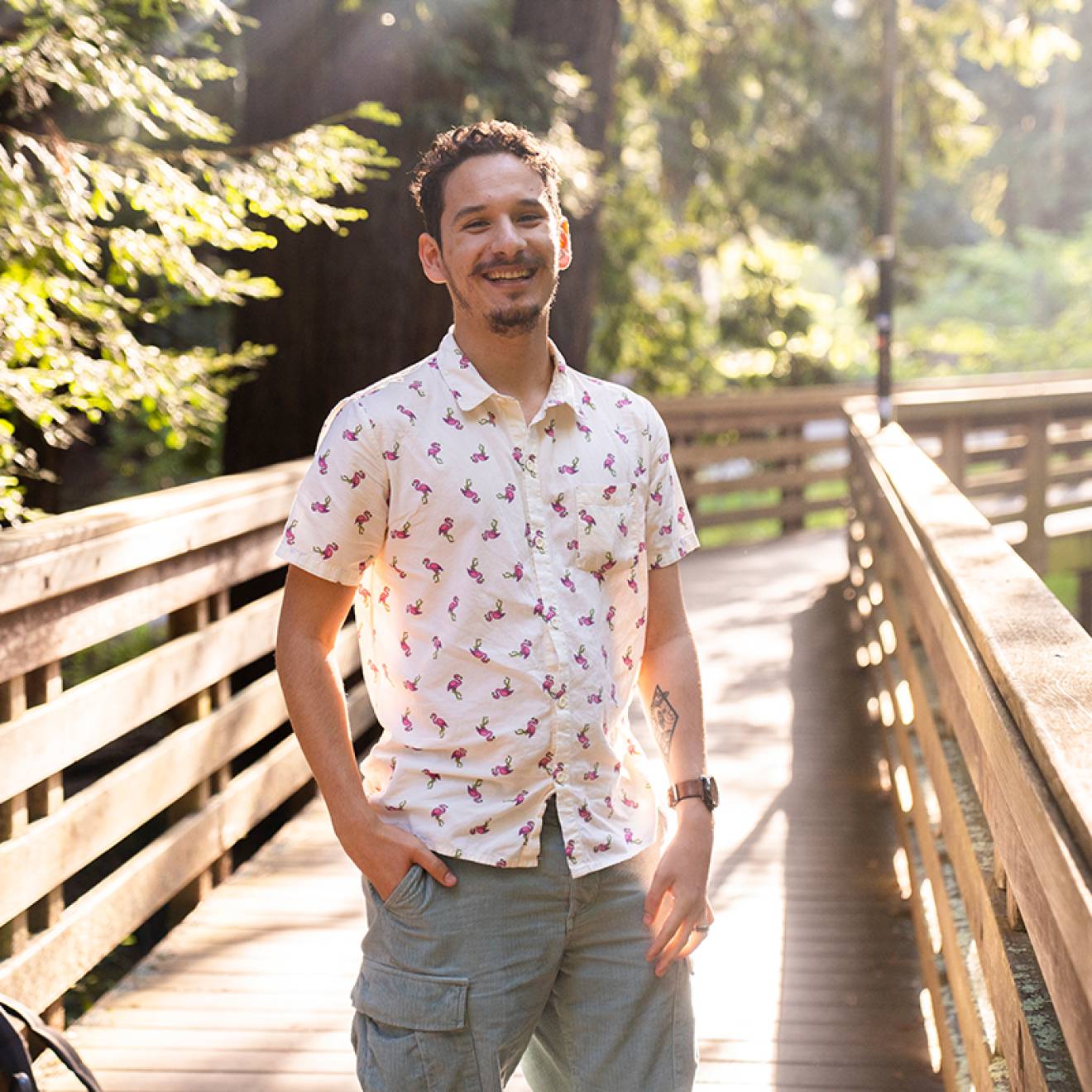 A smiling young man standing on a bridge surrounded by redwood trees