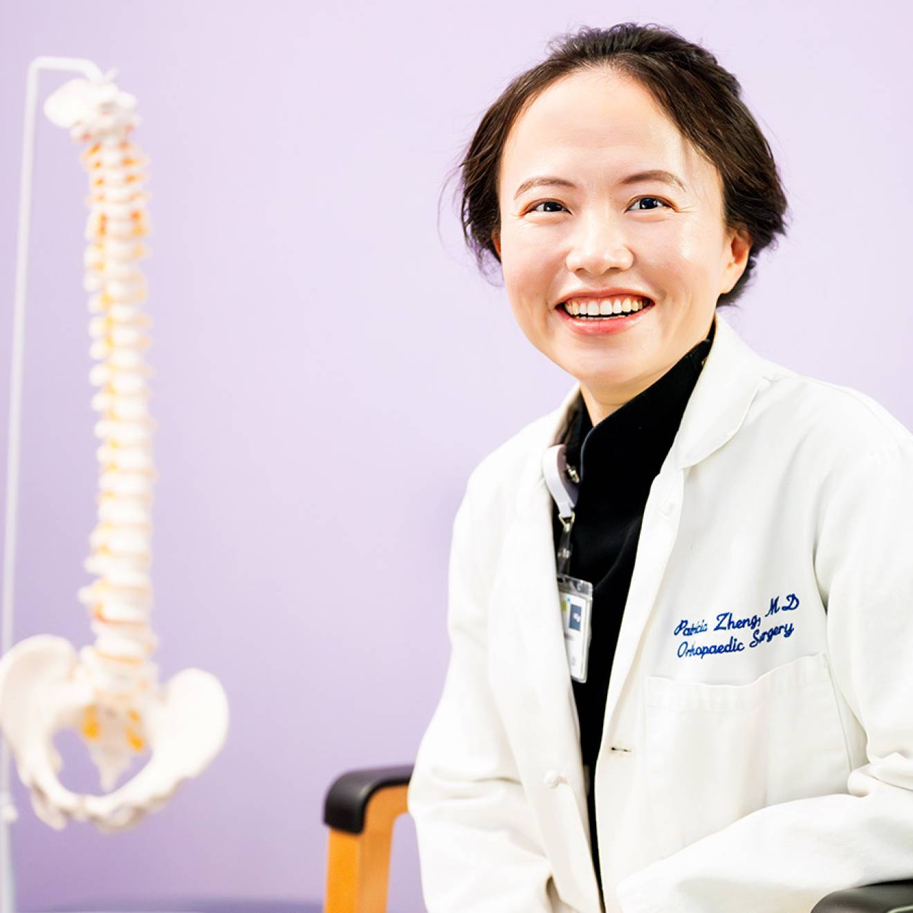 Spine health A woman with short hair in a white doctor's coat smiles in a chair with a model of a human spine behind her against a lilac backdrop