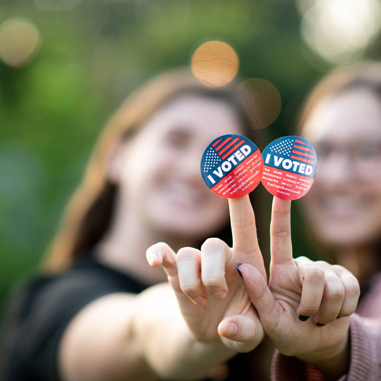Two young women, mostly out of focus, hold up I Voted stickers to the camera, smilling