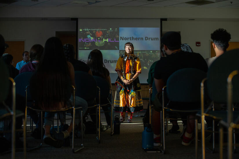 Young woman stands in front of darkened classroom in Native dress