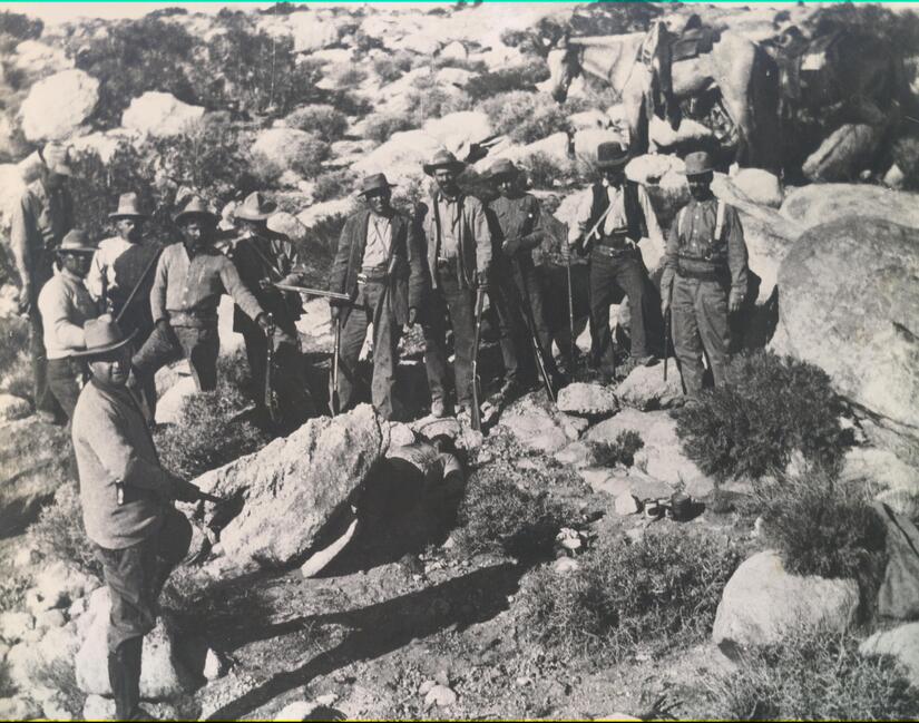 Old black and white photo of a group of men finding a body among rocks with its face obscured by shadow