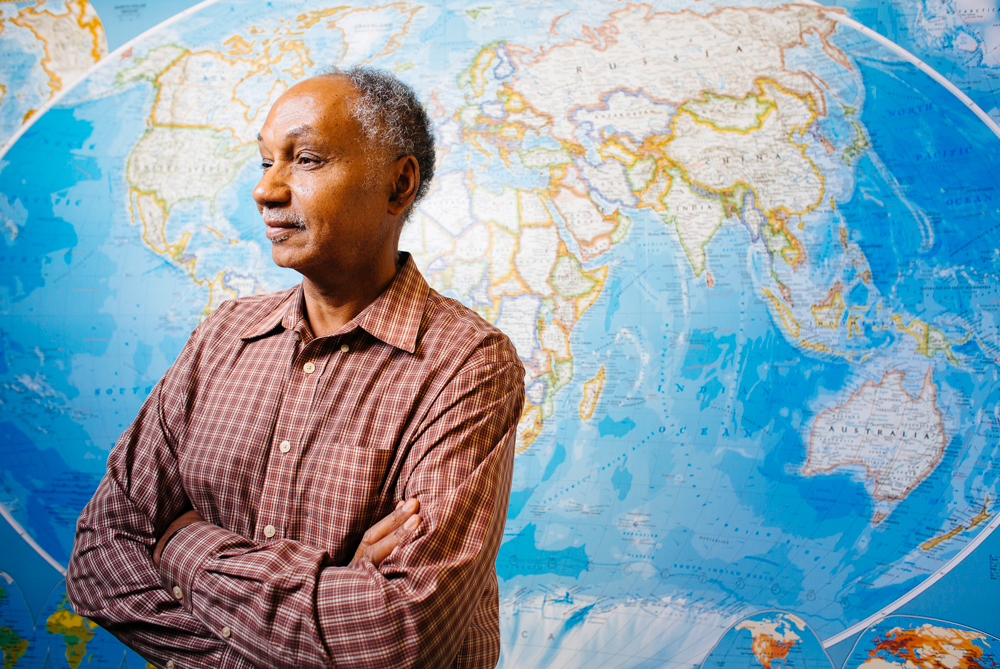 Older Black man standing in front of a map with arms crossed, smiling