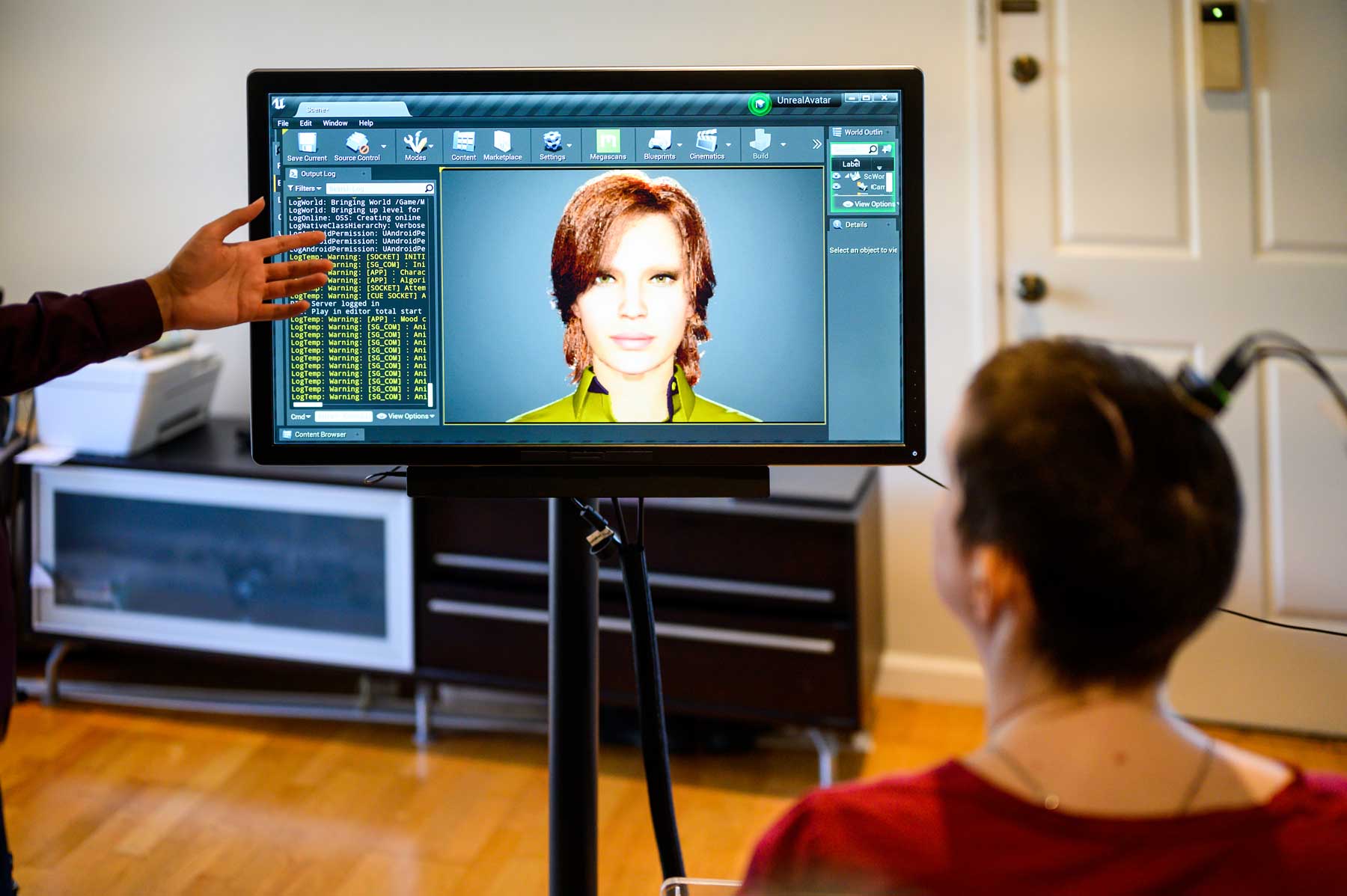 Ann, wearing an implant on her head, in red shirt, looks at a screen with a woman's face on it