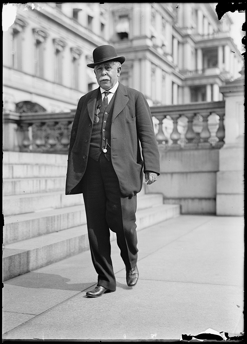Vintage black-and-white photo of a white-haired man in a three-piece suit and bowler hat in front of a Neoclassical building