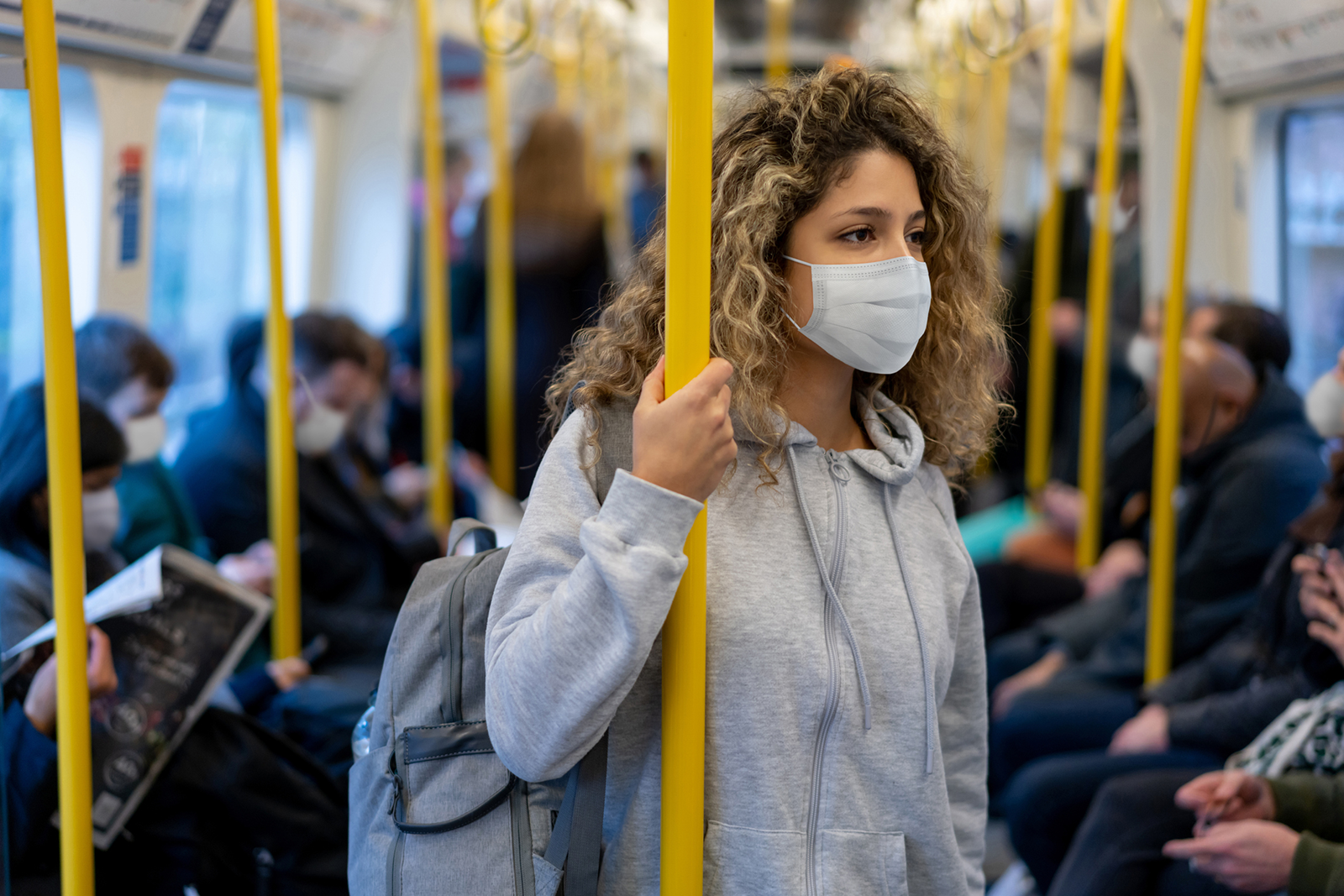 Young woman with curly hair wearing gray sweatshirt, backpack, and surgical face mask, stands on a crowded subway train holding on to a vertical yellow bar