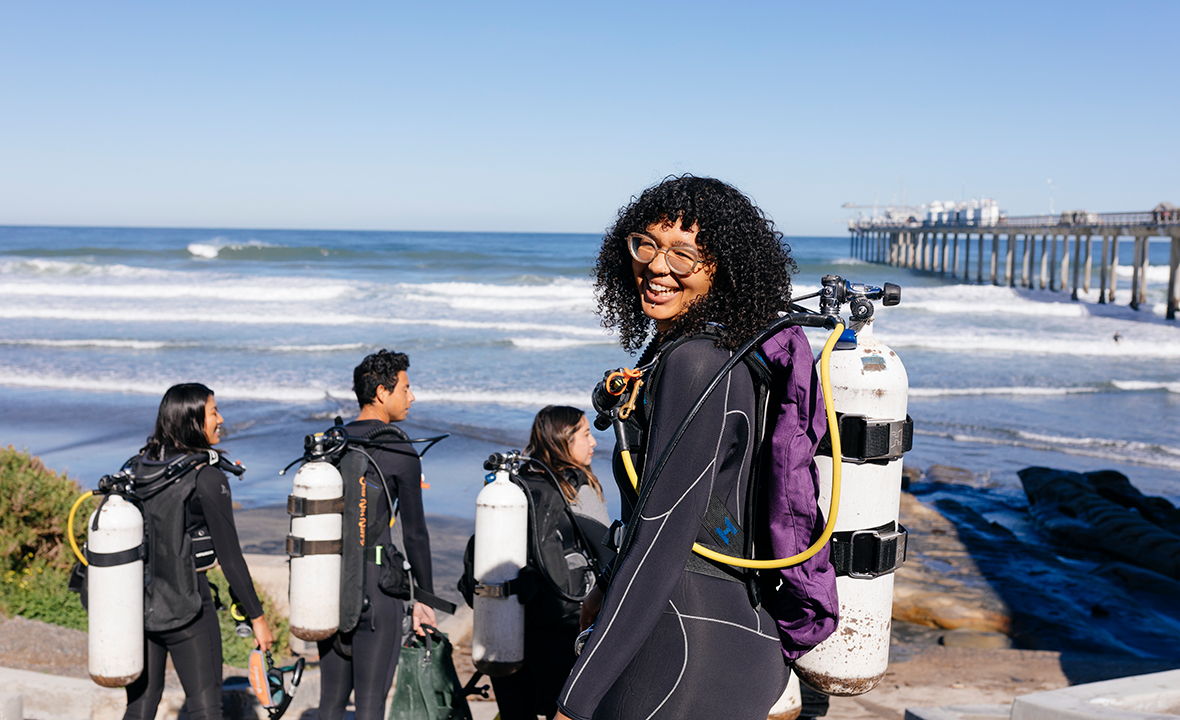 A group of students going out to scuba diva; a young African American woman smiling happily with a scuba tank on her back at the front of the photo