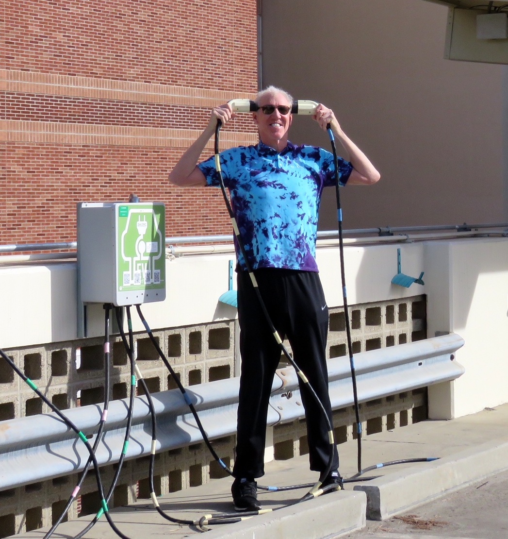 Bill Walton holds EV chargers up to his ears on the UCLA campus, wearing a tie-dyed shirt