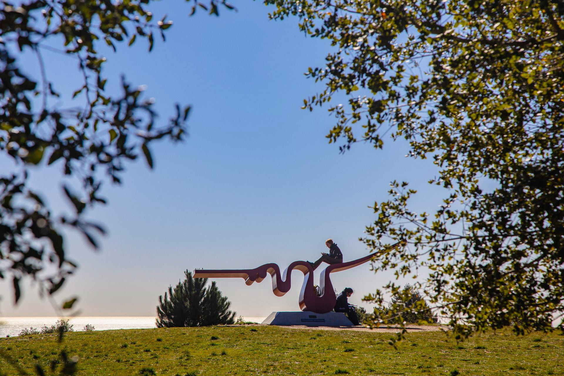 A person sitting on a large outdoor sculpture, seen through the trees with the ocean in the distance