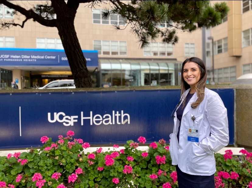 A young woman with long hair in a white coat stands next a UCSF Health sign outside, smiling