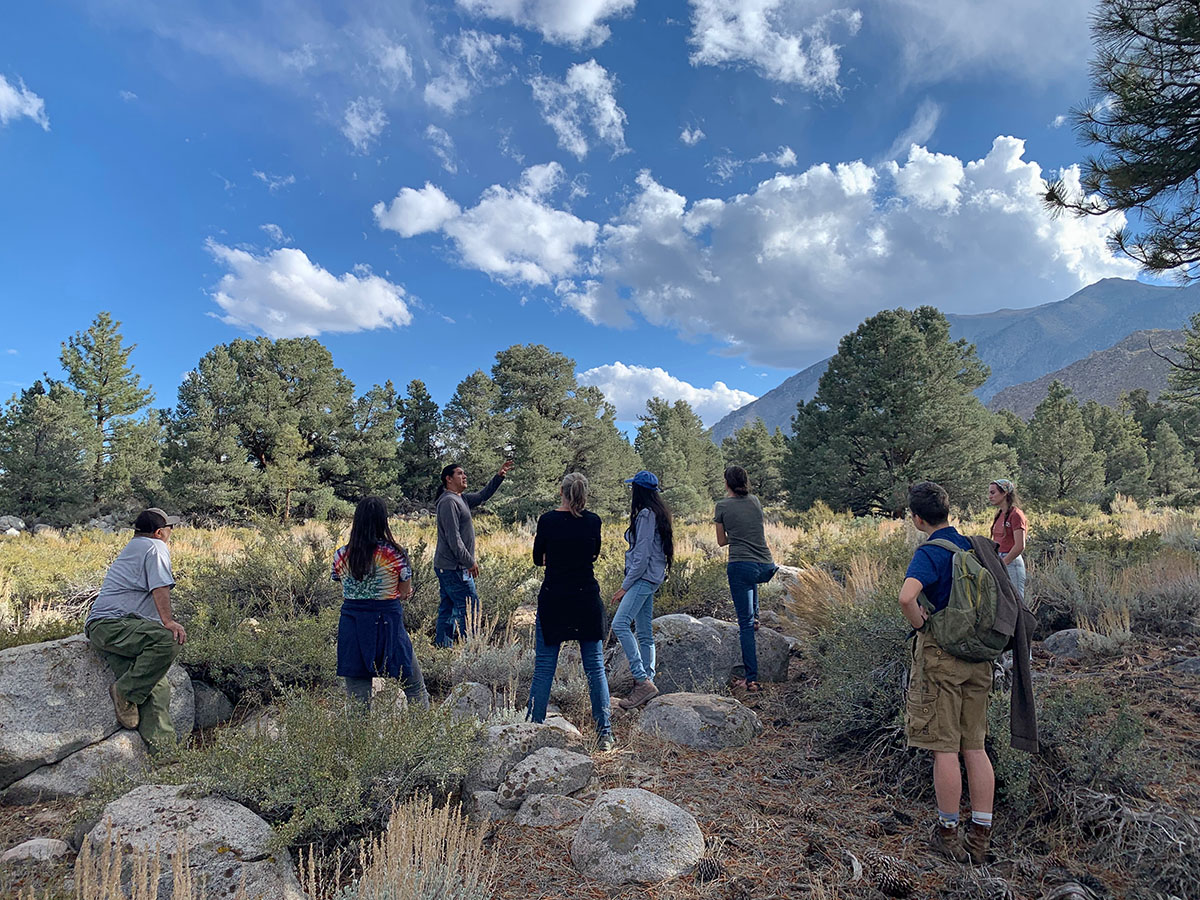 A group of people listed to a person speaking in a rocky and shrubby area with pinyon pines just beyond