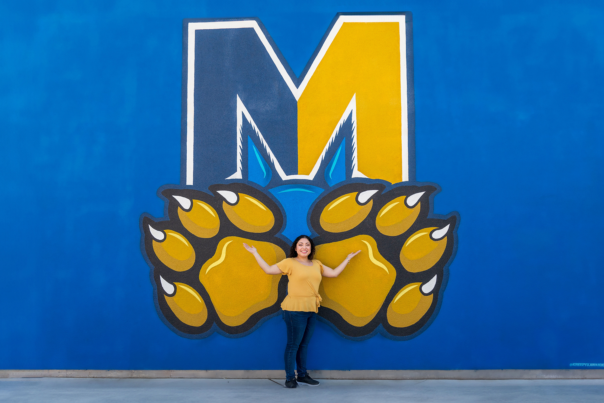 A woman stands in front of the Merced "M" with hands extended over the bobcat paws