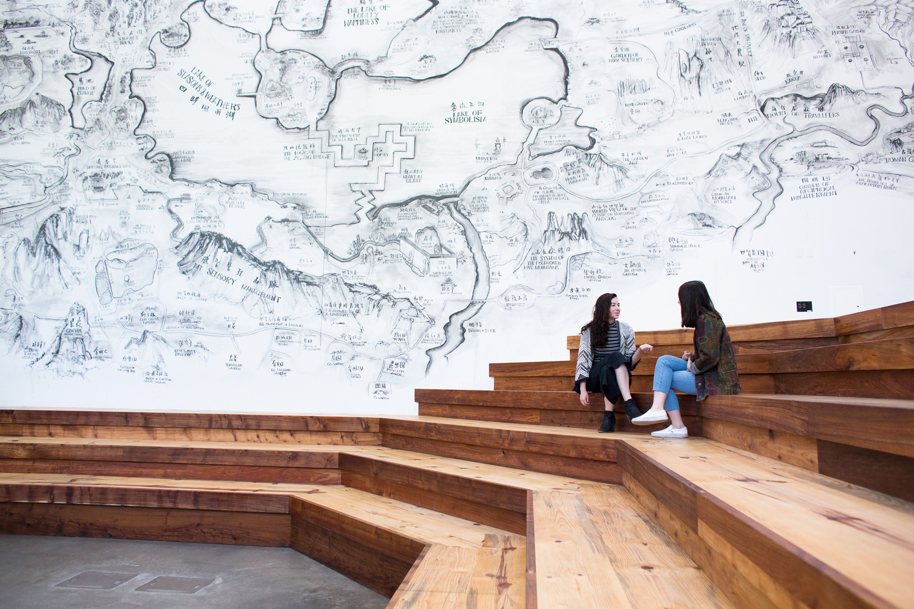 Two people in conversation sitting on wood steps in front of an indoor mural