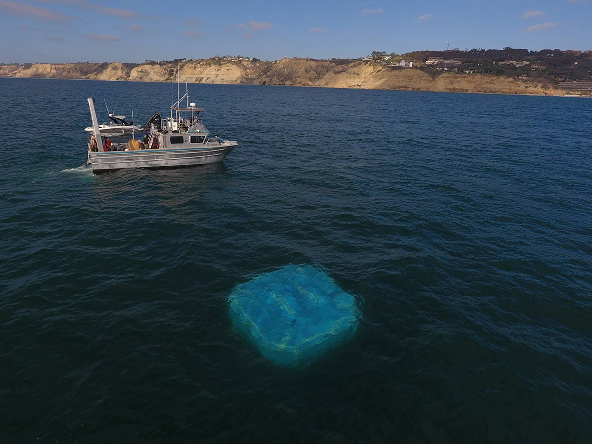 Blue platform floats beneath ocean surface, attended by silver fishing boat, a few hundred meters offshore
