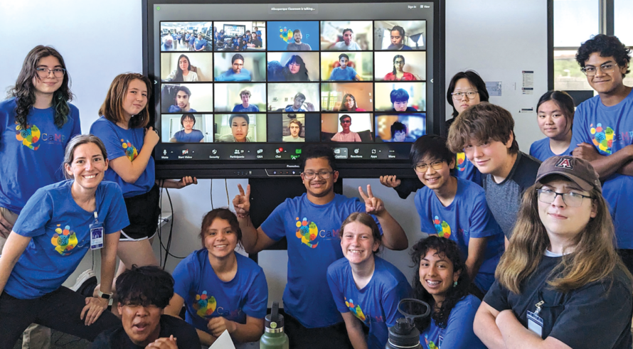 A group of preteens and a few adults in matching blue T-shirts surround people dressed similarly on a large screen on Zoom