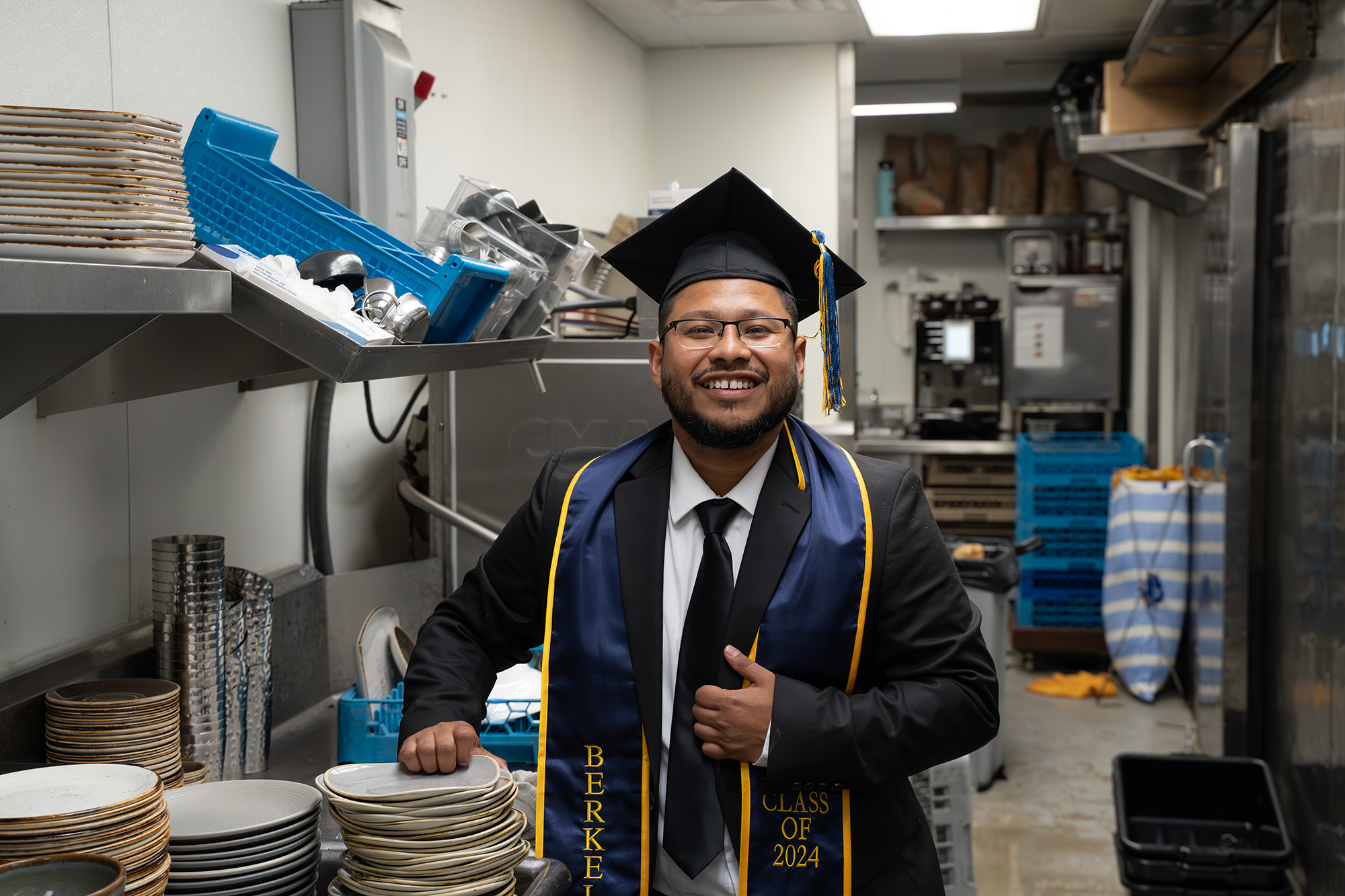 A bearded graduate in cap, gown and stole poses, smiling, in a commercial kitchen