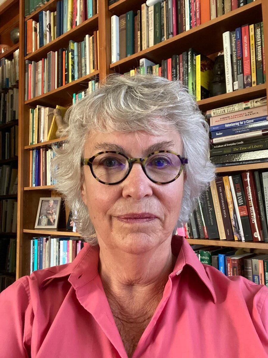 Woman with gray hair and oval glasses in pink shirt in front of a wall of books