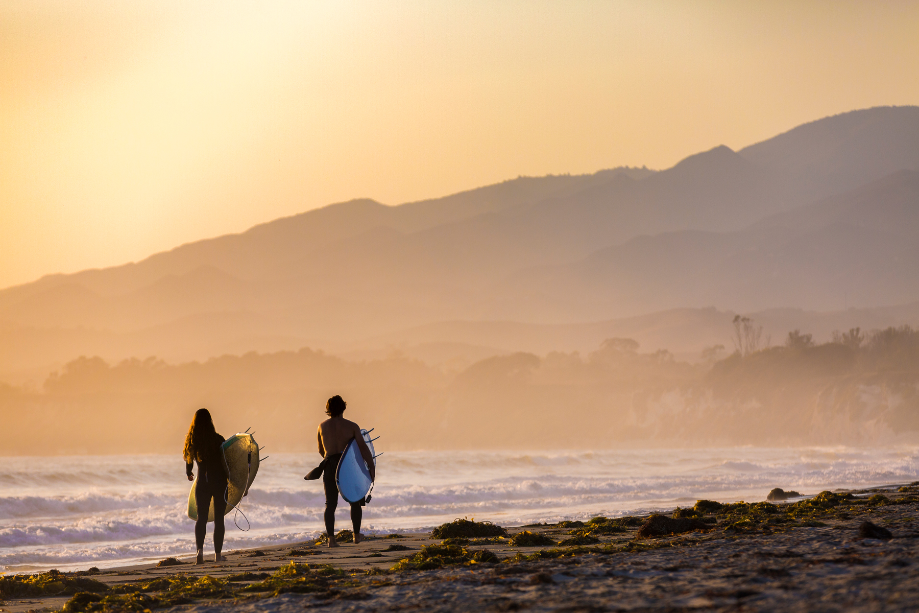 Two surfers walking along the beach at sunset with mountains in the background