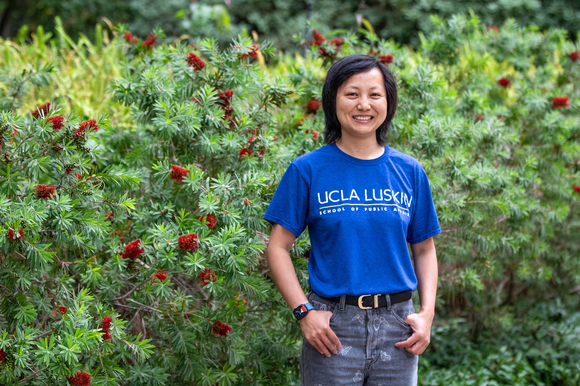 Woman in blue UCLA Luskin s hirt and jeans stands, smiling, in front of a green bush with red blooms