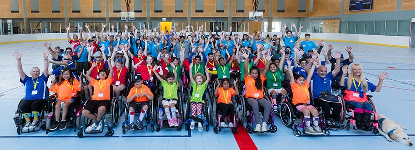 A large group photo of people in wheelchairs with their arms raised, colorful T-shirts worn, a yellow lab next to a person on the far right