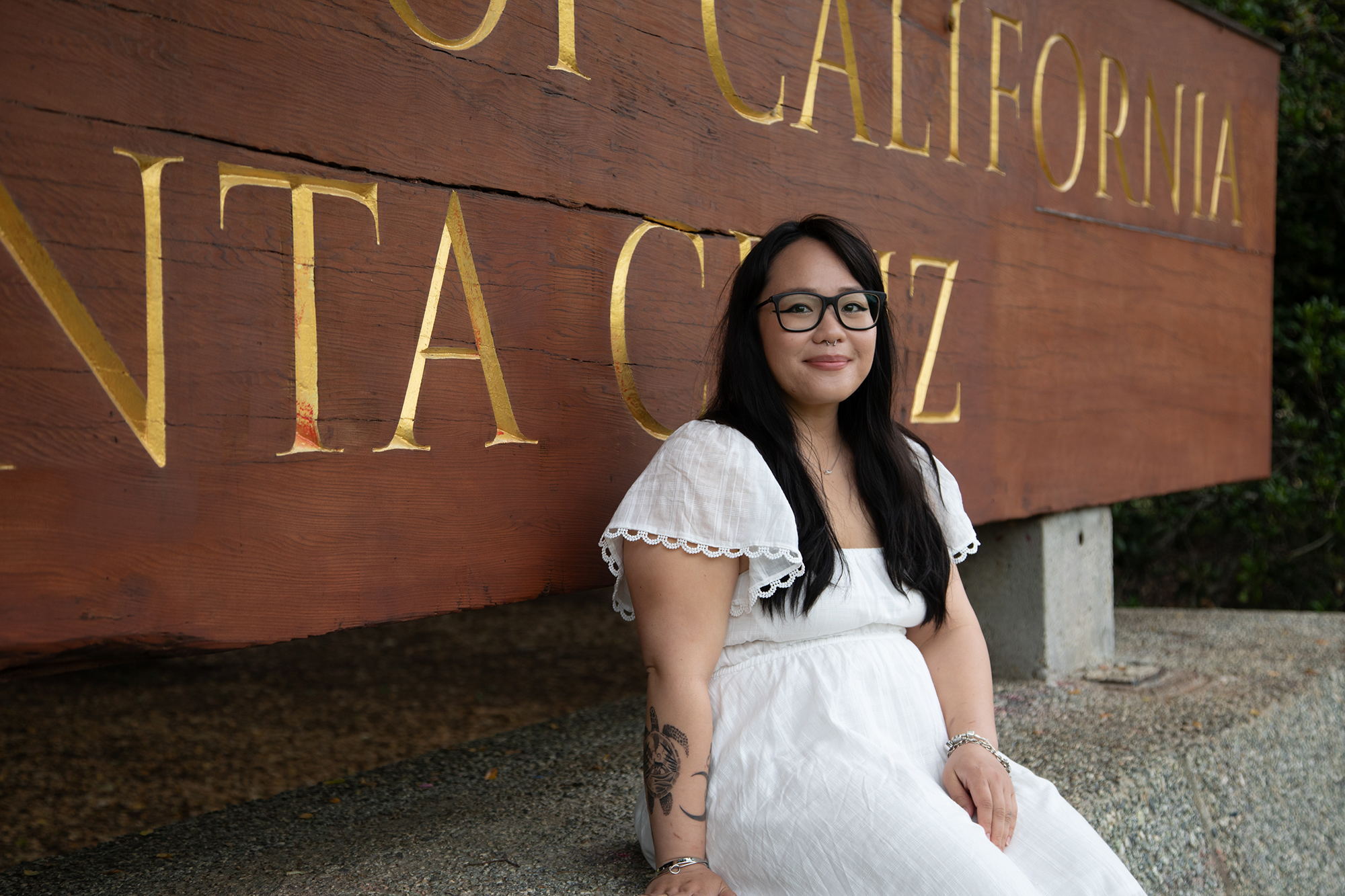 Young woman with long hair, glasses and white dress sits in front of a large wooden University of California Santa Cruz sign