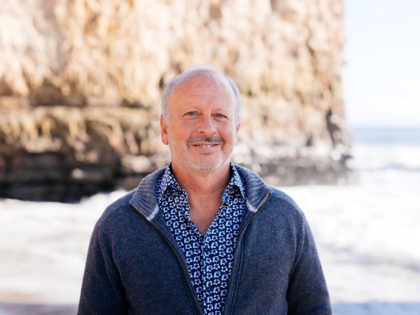 Mike Beck smiles on a beach with a cliff in the background
