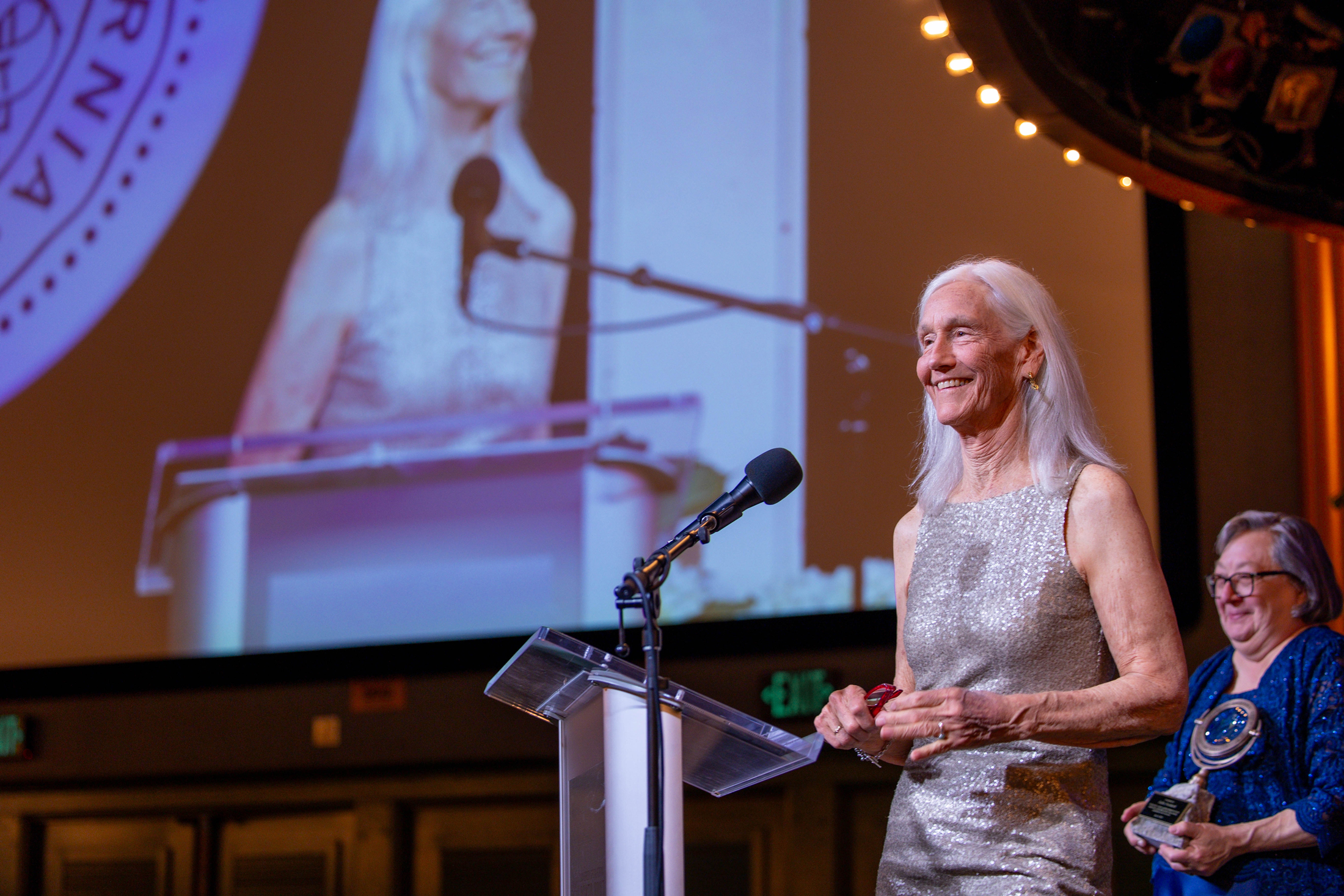 Julie Packard, woman with long white hair in a silvery gown, speaks at a podium, a screen showing her image behind her, with UC Santa Cruz Chancellor Cynthia Larive, in short gray hair and a blue dress, looks on