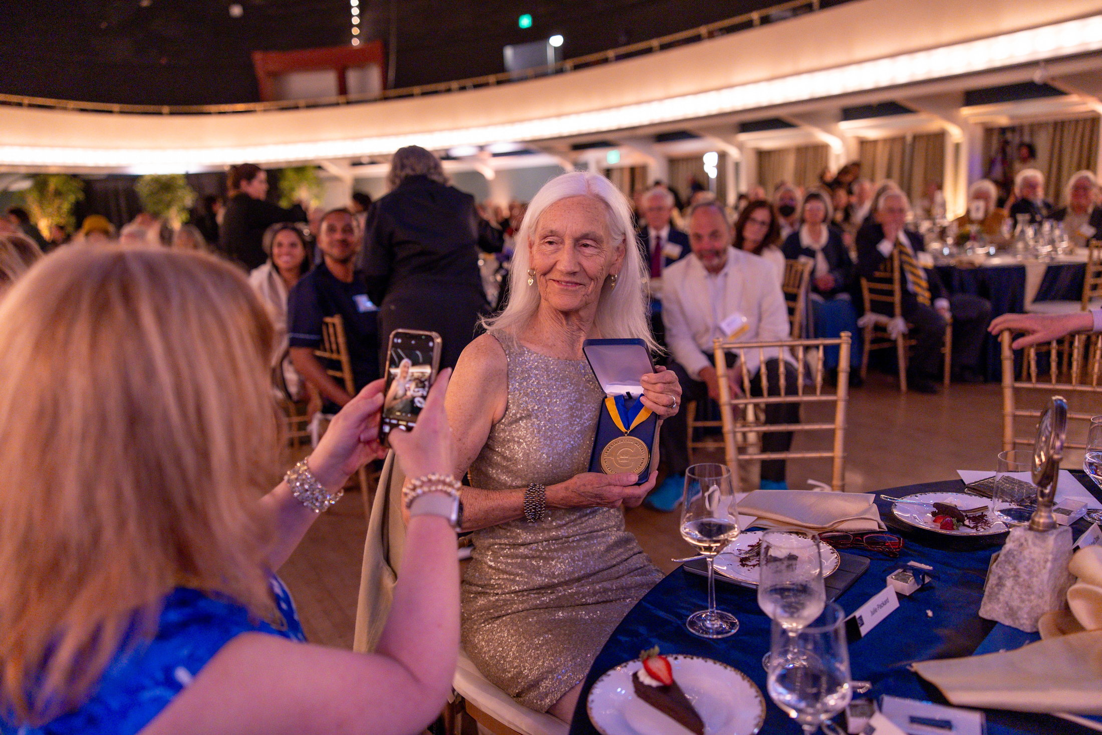 Julie Packard displays the UC Presidential Medal at a dinner table in a crowded banquet hall