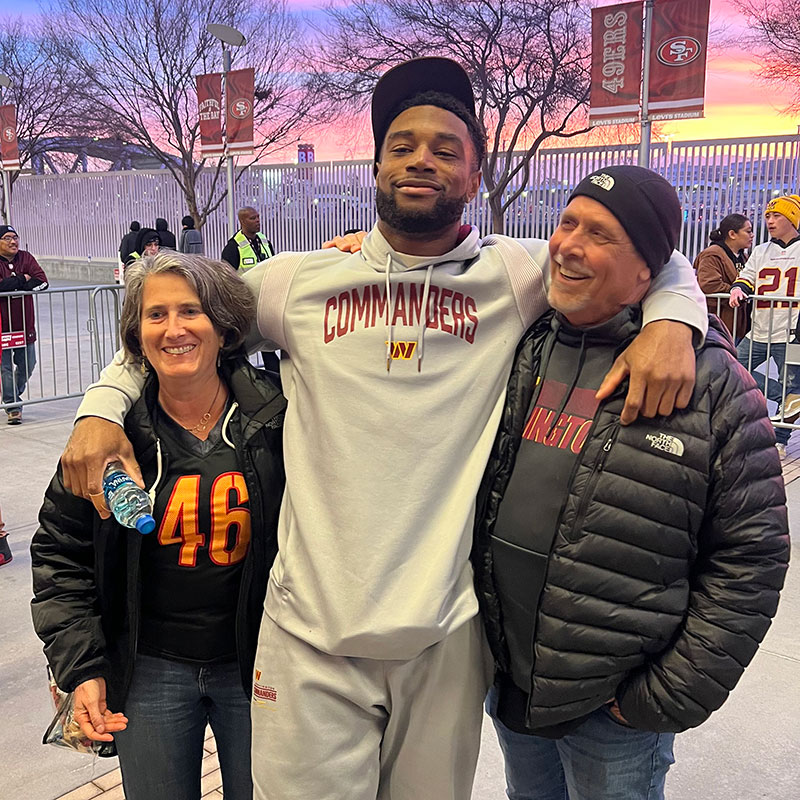 A young man wearing a sports sweatshirt stands with his arms around his parents' shoulders 