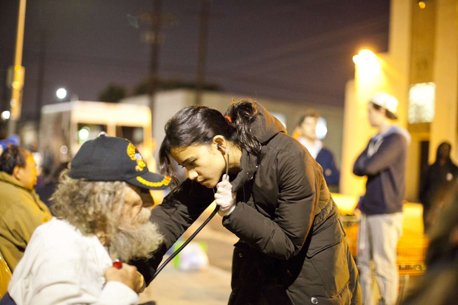 A student wearing a puffy coat uses a stethoscope to examine a gentleman with unkempt hair and beared wearing a military veterans hat, outdoors at night.