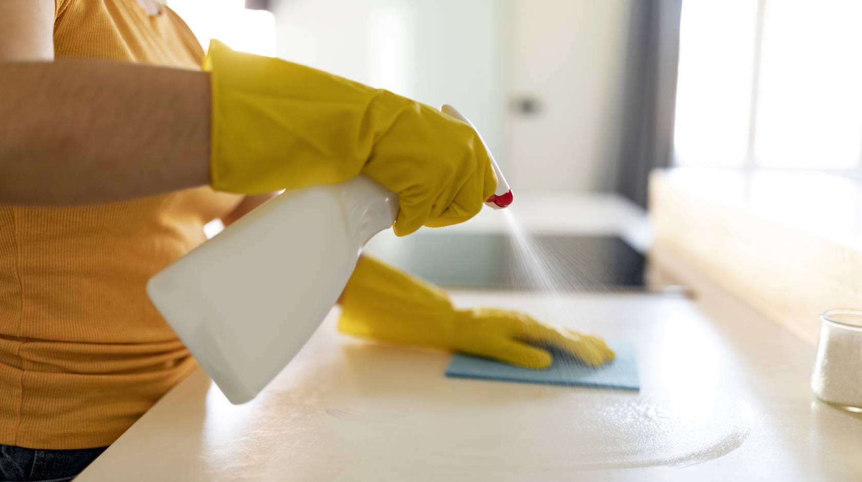 Person wearing yellow rubber gloves sprays cleaner on a countertop and wipes it with a sponge