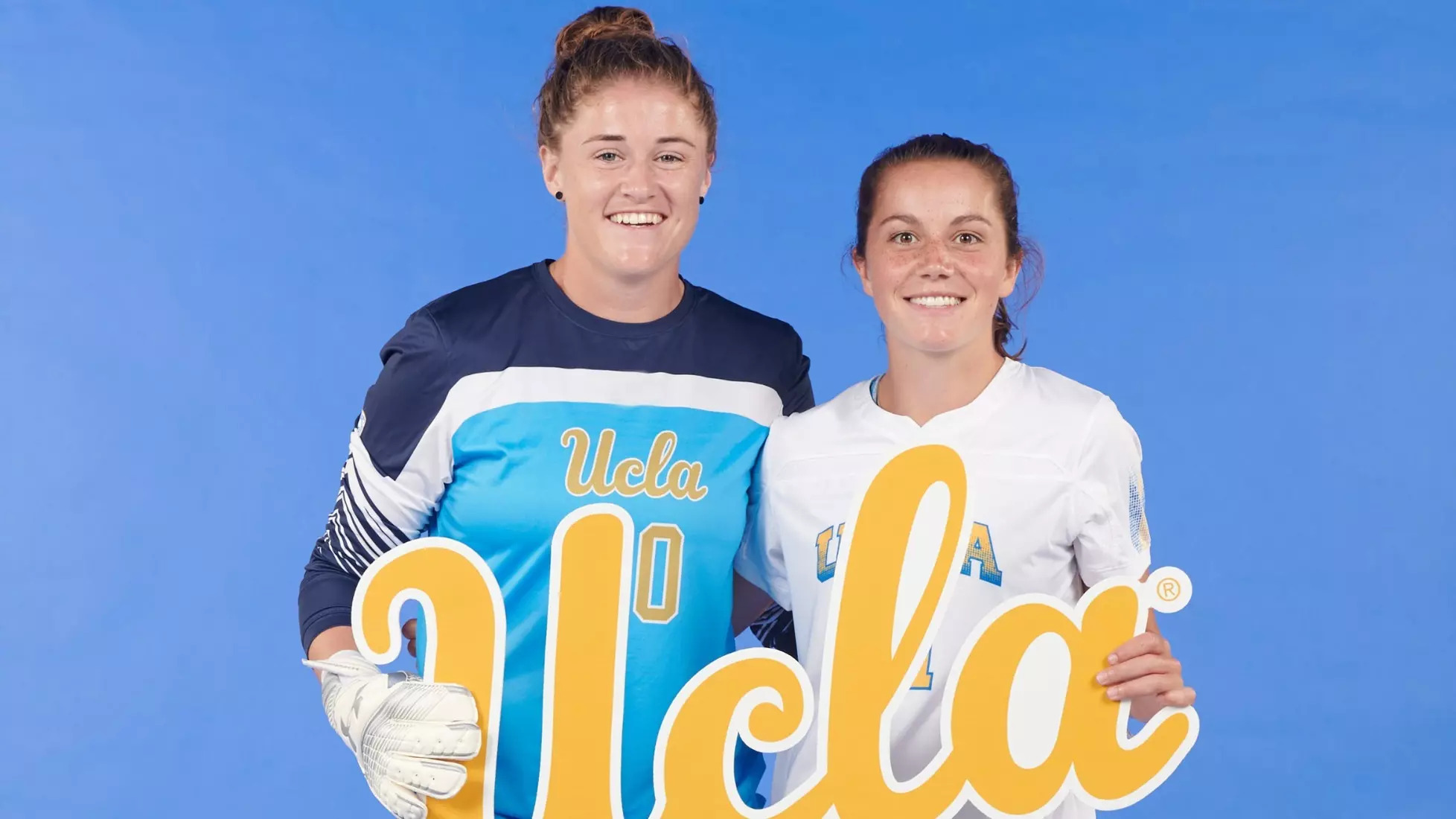 Two young women, on the left, Teagan Micah wearing a goalkeeper's gloves and uniform, on the right, Jessie Fleming with a field player uniform, holding a UCLA sign and smiling