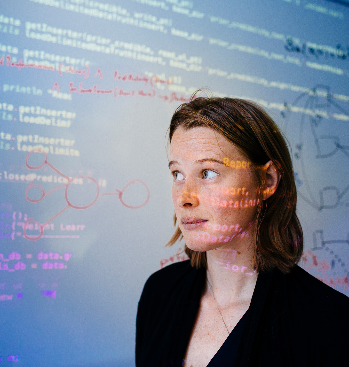 A young woman quarter-faces the camera, looking up and to the right. She's standing in front of a white board with some technical drawings, and computer code is projected on the white board and onto her face. 