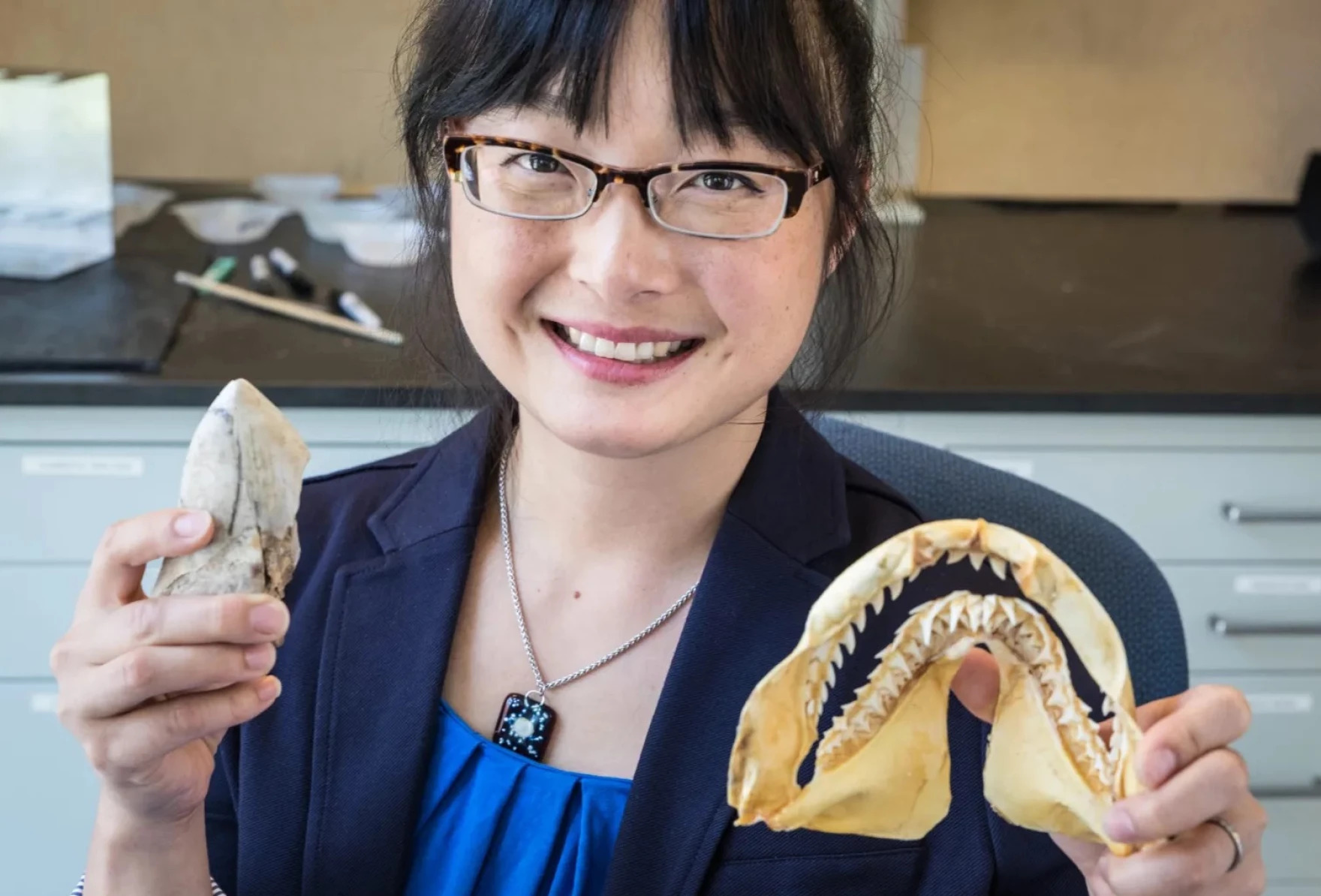 Smiling woman with glasses holds up shark teeth and jaw
