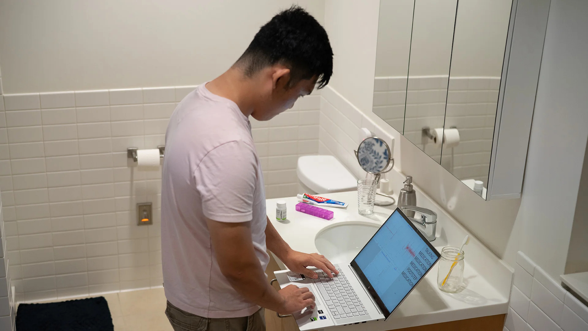 A man typing on a laptop set up on a bathroom sink