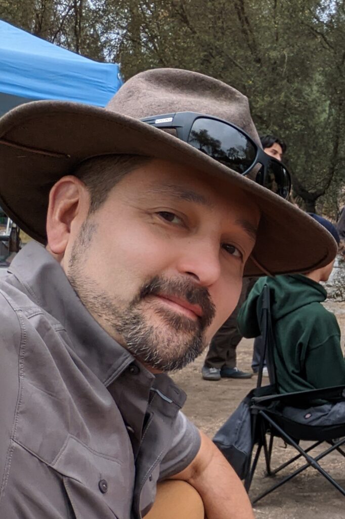 Jun Sunseri, wearing an Indiana Jones-style hat, leans forward and turns his head to the right to smile at the camera while seated at a campground