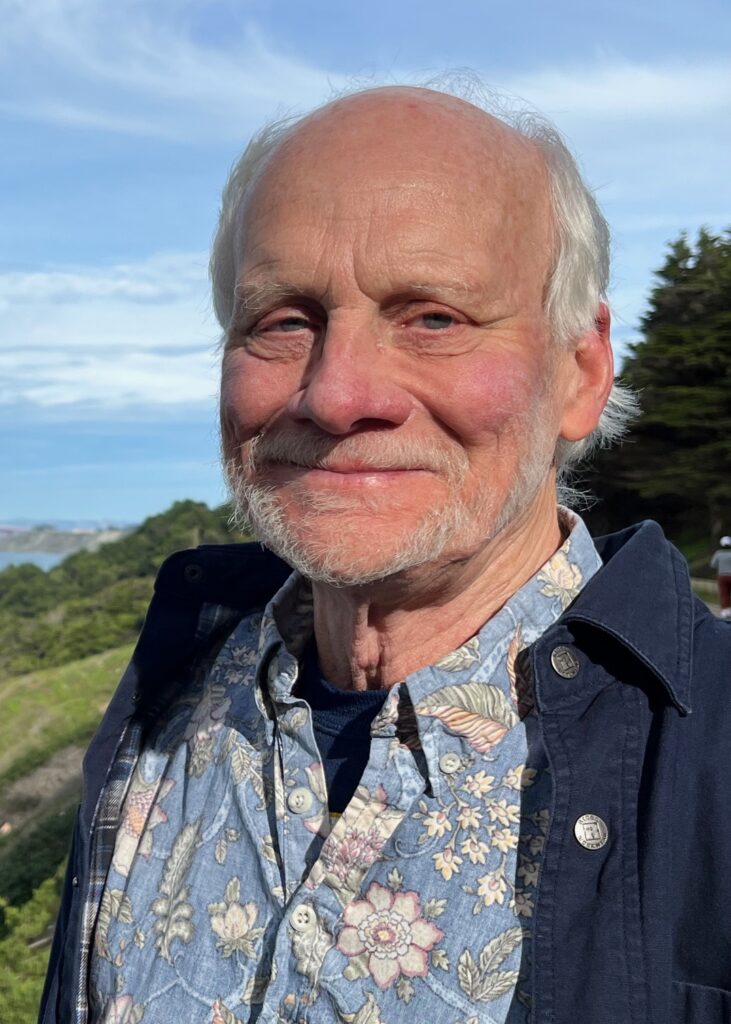 Kent Lightfood smiles at the camera wearing a floral shirt and jacket, outside on a green hillside