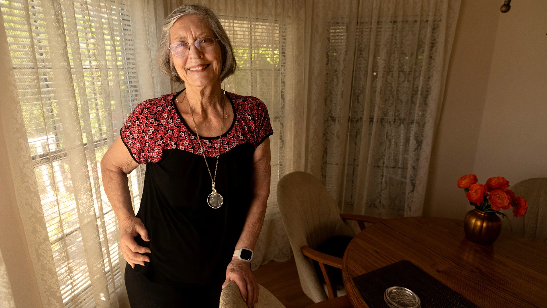 A woman with gray hair standing in her dining room with lace curtains in the background