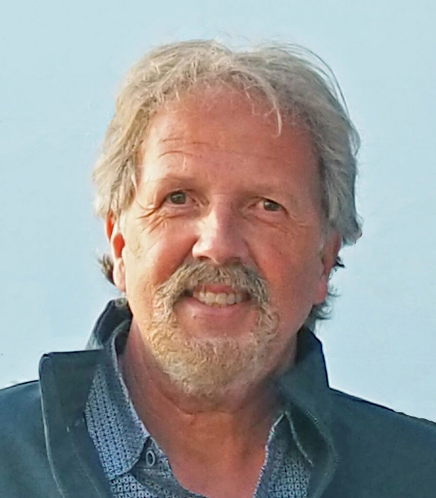 Scott Byram smiles in a headshot, outdoors against blue sky, in a blue button up and dark blue jacket.