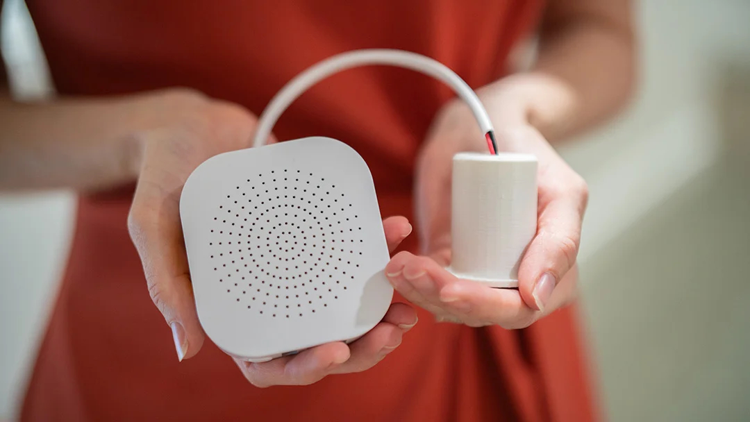 A palm-sized white plastic square box with rounded corners and holes in a circular pattern, connected by a wire to a smaller cylindrical box.