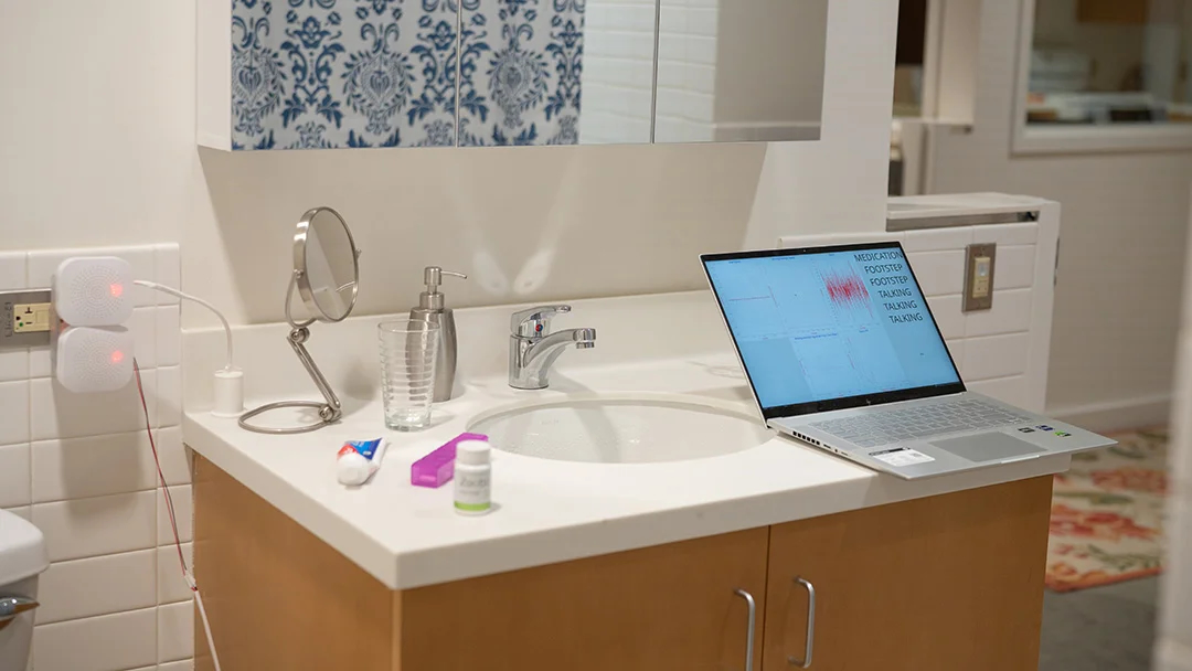 A bathroom sink with a laptop computer sitting on the edge of the vanity and a sensor plugged into the wall behind it