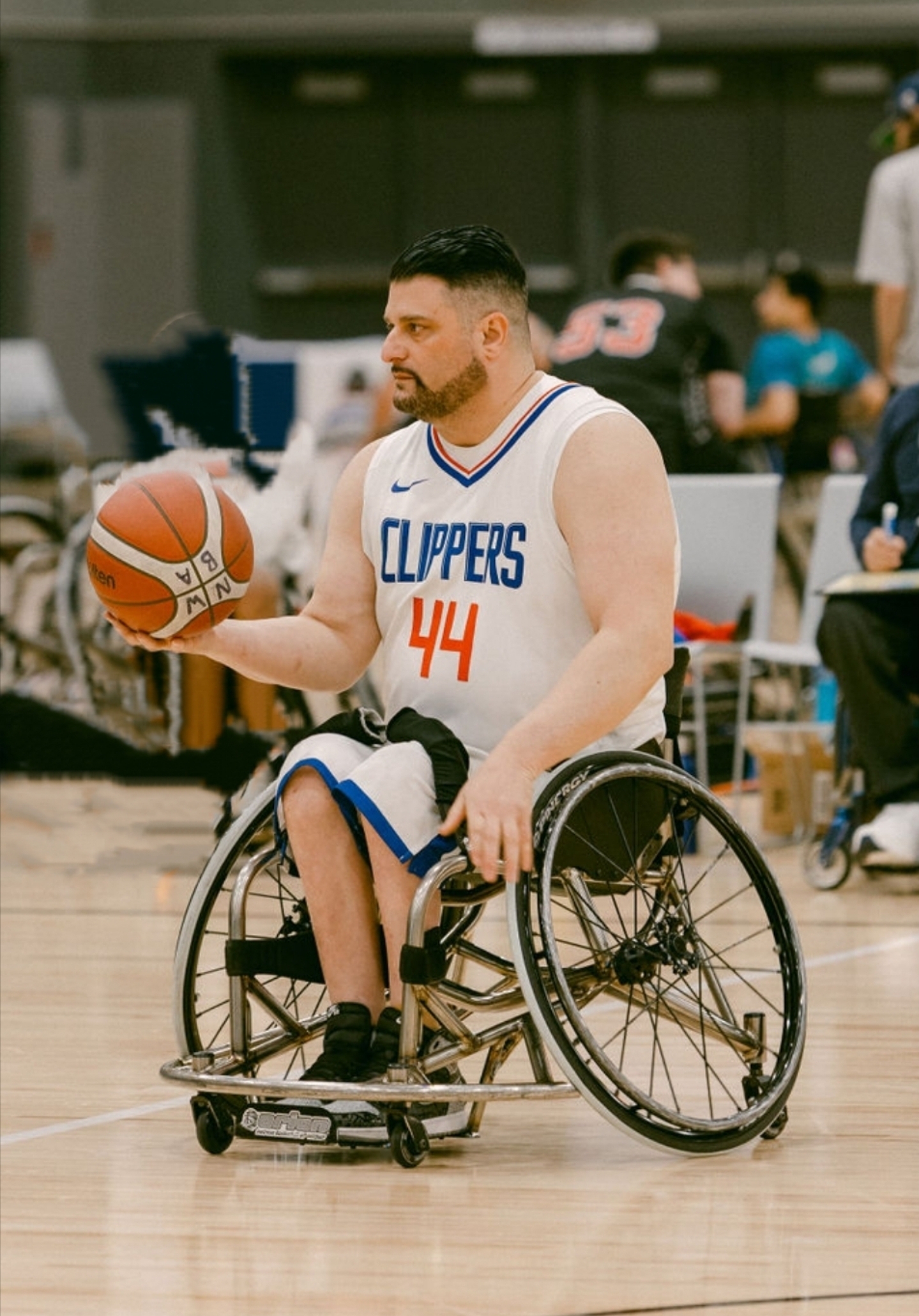 Man with a beard in a wheelchair holding a basketball on a court, wearing a LA Clippers jersey with the number 44